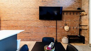 A brick-walled room with a flat-screen TV mounted on the wall, a black table with chairs, and a small shelf with hats and a basket. - Retro Vibe,  downtown Grand Forks (Grand Forks)