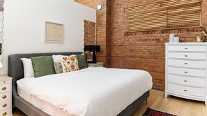 A bedroom with a brick wall, a bed with a white comforter, a white dresser, and a bedside table with a lamp. - Retro Vibe,  downtown Grand Forks (Grand Forks)