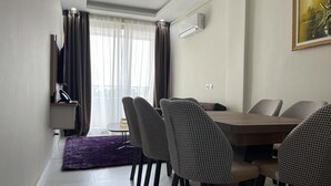 A modern dining area with a wooden table and houndstooth chairs, a purple rug, and a window with curtains. - RESIDENCE SYCOMORE 1 (Abidjan)