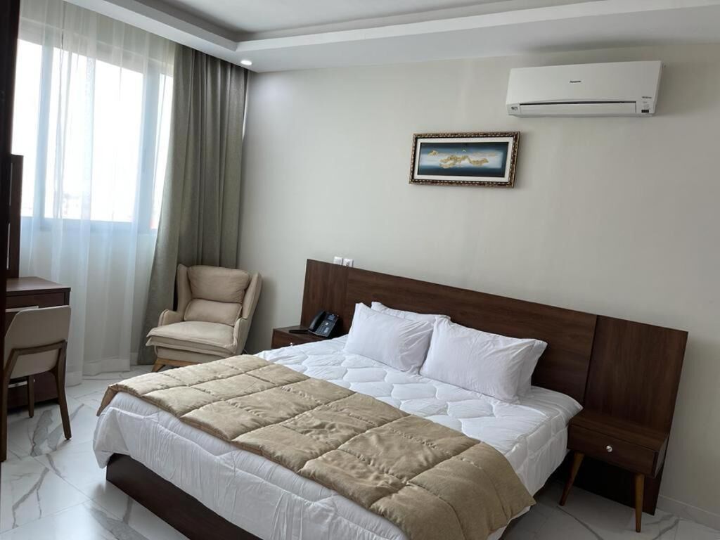 A hotel room with a large bed, a chair, a desk, and an air conditioning unit.