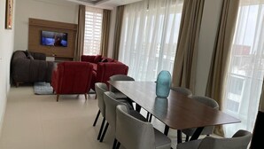 A modern living room with a dining area, a flat-screen TV mounted on the wall, and large windows with sheer curtains. - RESIDENCE SYCOMORE 1 (Abidjan)