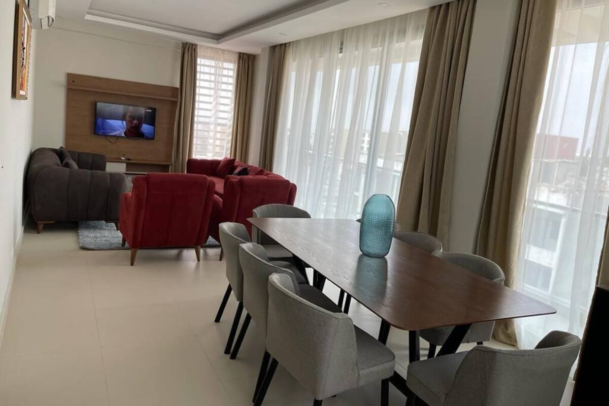 A modern living room with a dining area, a flat-screen TV mounted on the wall, and large windows with sheer curtains.