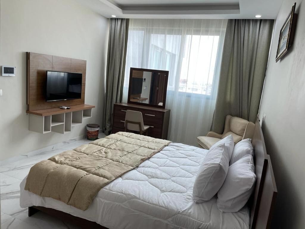 Habitación de hotel con cama, televisor montado en un panel de madera, escritorio con silla y una ventana grande con cortinas.