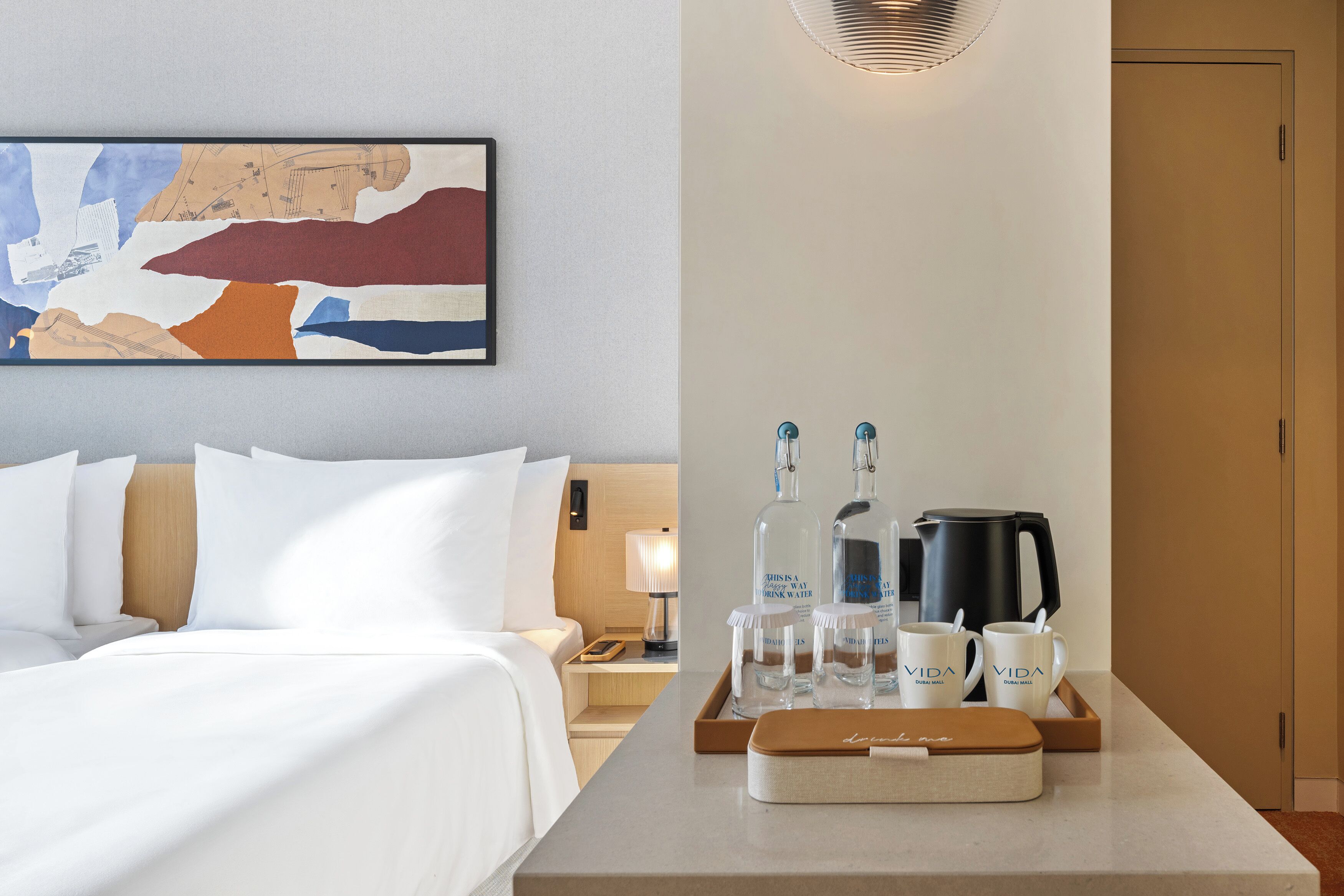 Habitación de hotel con una cama bien tendida, una bandeja con botellas y vasos, y un cuadro en la pared.