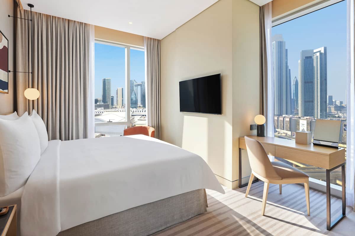 A hotel room with a large bed, a desk, a chair, a TV, and a view of the cityscape.