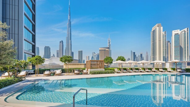 Una piscina con sillones de descanso y sombrillas, rodeada de edificios modernos y un cielo despejado.