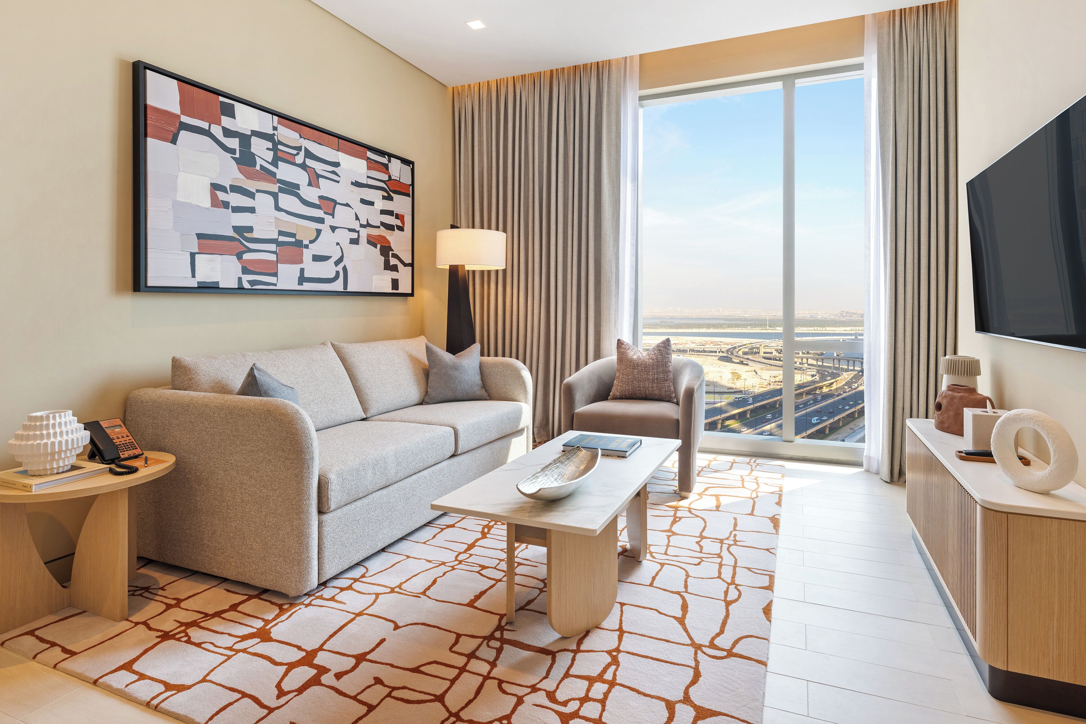 Une chambre d’hôtel moderne, dotée d’un canapé, d’un fauteuil et d’une table basse. Une grande fenêtre offre une vue sur le paysage urbain.