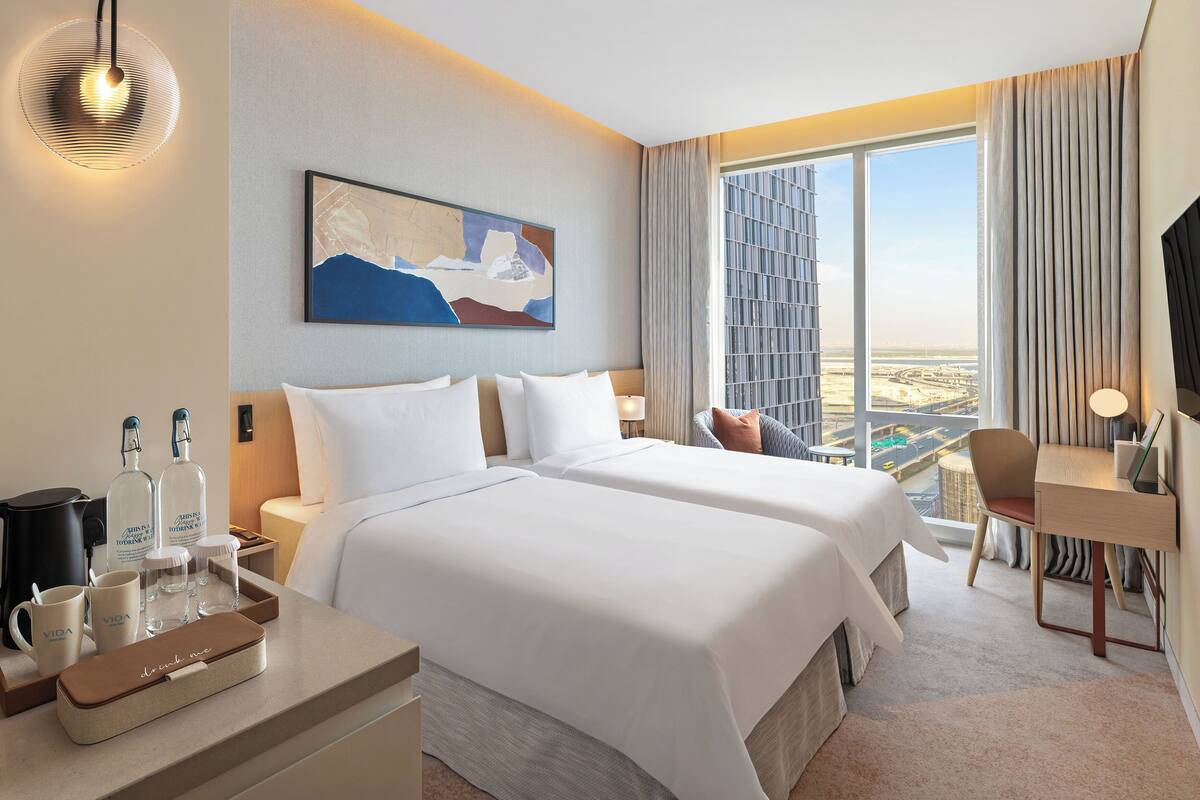 A hotel room with two beds, a desk, a chair, a TV, a painting, and a view of a cityscape.
