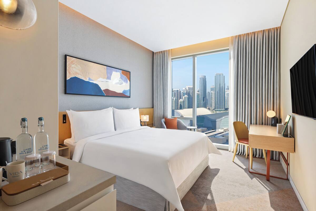 A hotel room with a large bed, a desk, a chair, a TV, and a city view.