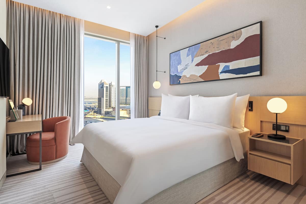A modern hotel room with a large bed, a desk, a chair, and a city view.