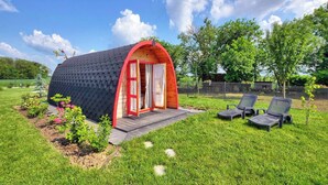 Una cabaña pequeña de una sola habitación, con puerta roja y techo curvo, rodeada por un césped y dos sillones de descanso.