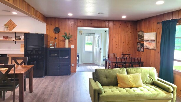 A cozy living room with a green sofa, wooden walls, and a dining area with a table and chairs. - Walk to the Concerts! (Arnolds Park)