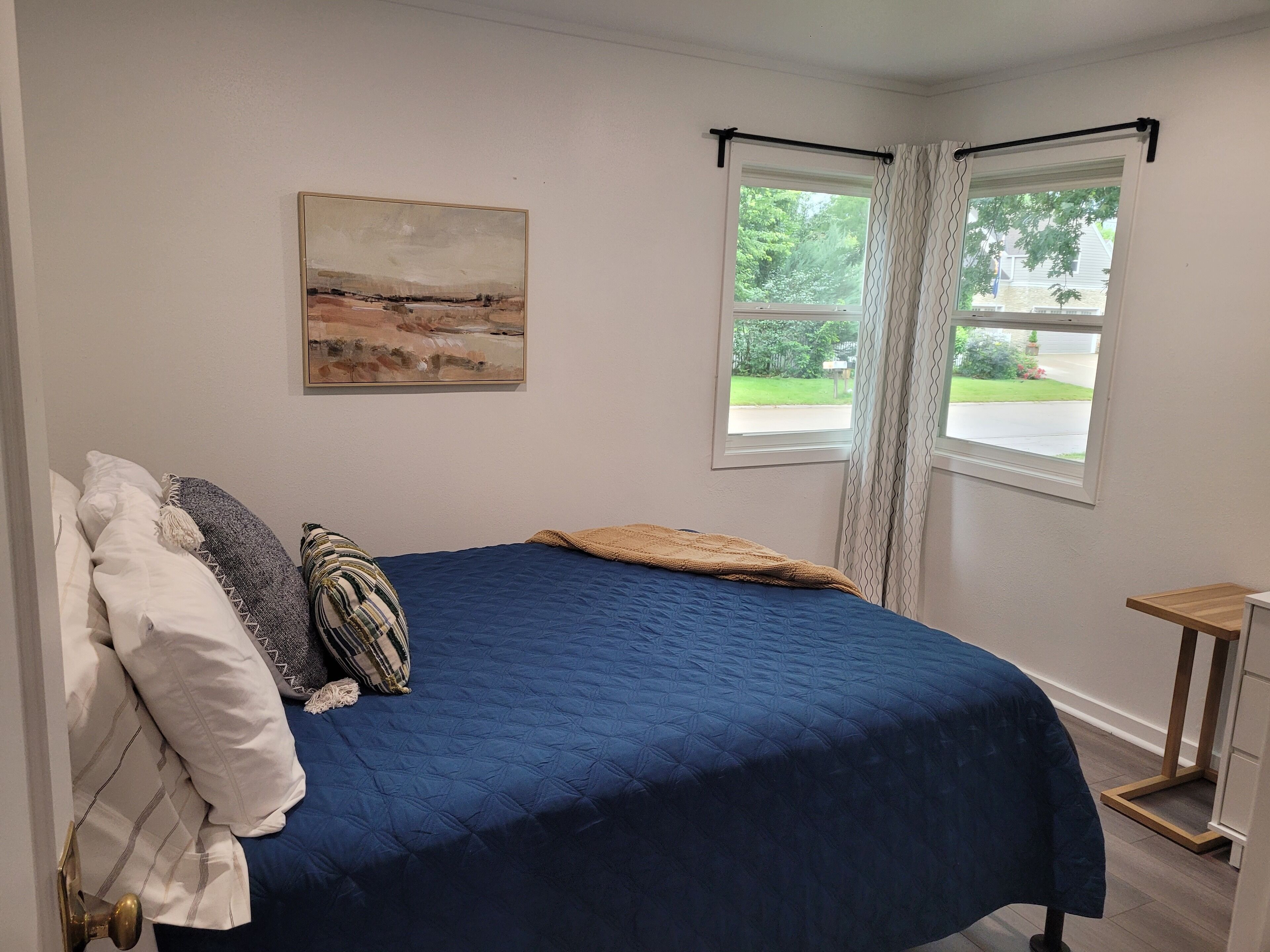 Un dormitorio con cama, ventana con cortinas, una mesita y un cuadro en la pared.