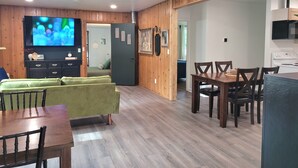 A modern living room with a green sofa, wooden dining table, and chairs, a flat-screen TV, and a kitchen area with a stove. - Walk to the Concerts! (Arnolds Park)