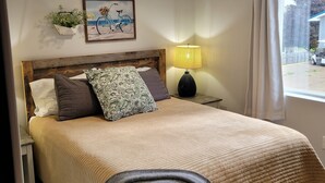 Een net opgemaakt bed met een beddengoed met patroon, een hoofdbord met een rustieke afwerking, een aan de muur bevestigde plant, een lijst met een afbeelding van een fiets en een bedlampje.