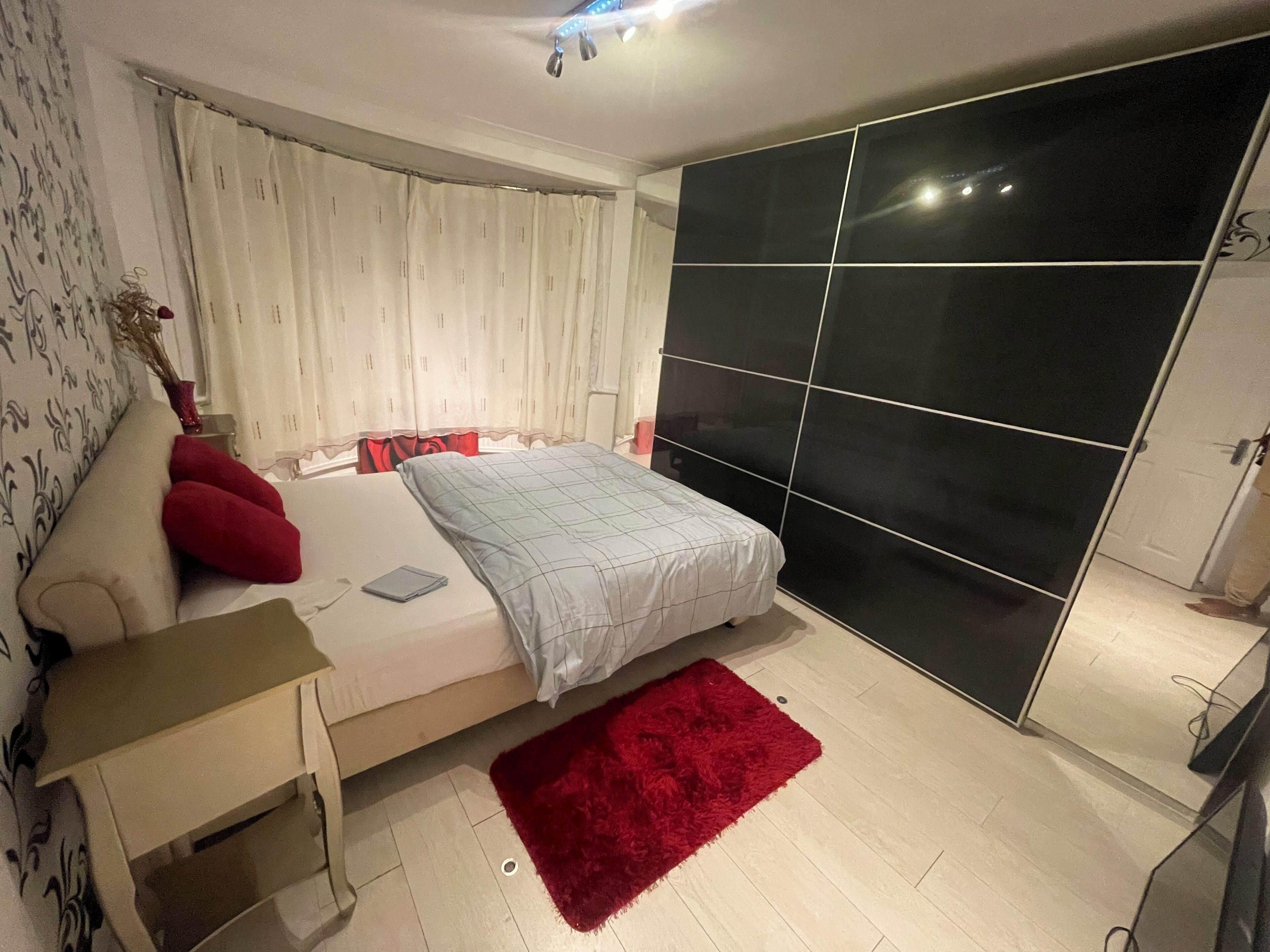 A bedroom with a bed, a nightstand, a red rug, a white curtain, and a black partition wall.
