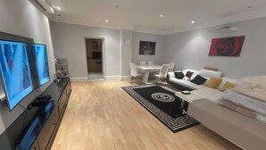 A modern living room with a flat-screen TV, a sofa, a dining table, and a kitchen area in the background.