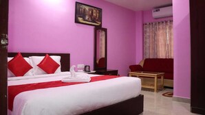 A hotel room with a bed, a red sofa, a small table, and a painting on the wall. - Hotel AMR GRAND (Port Blair)