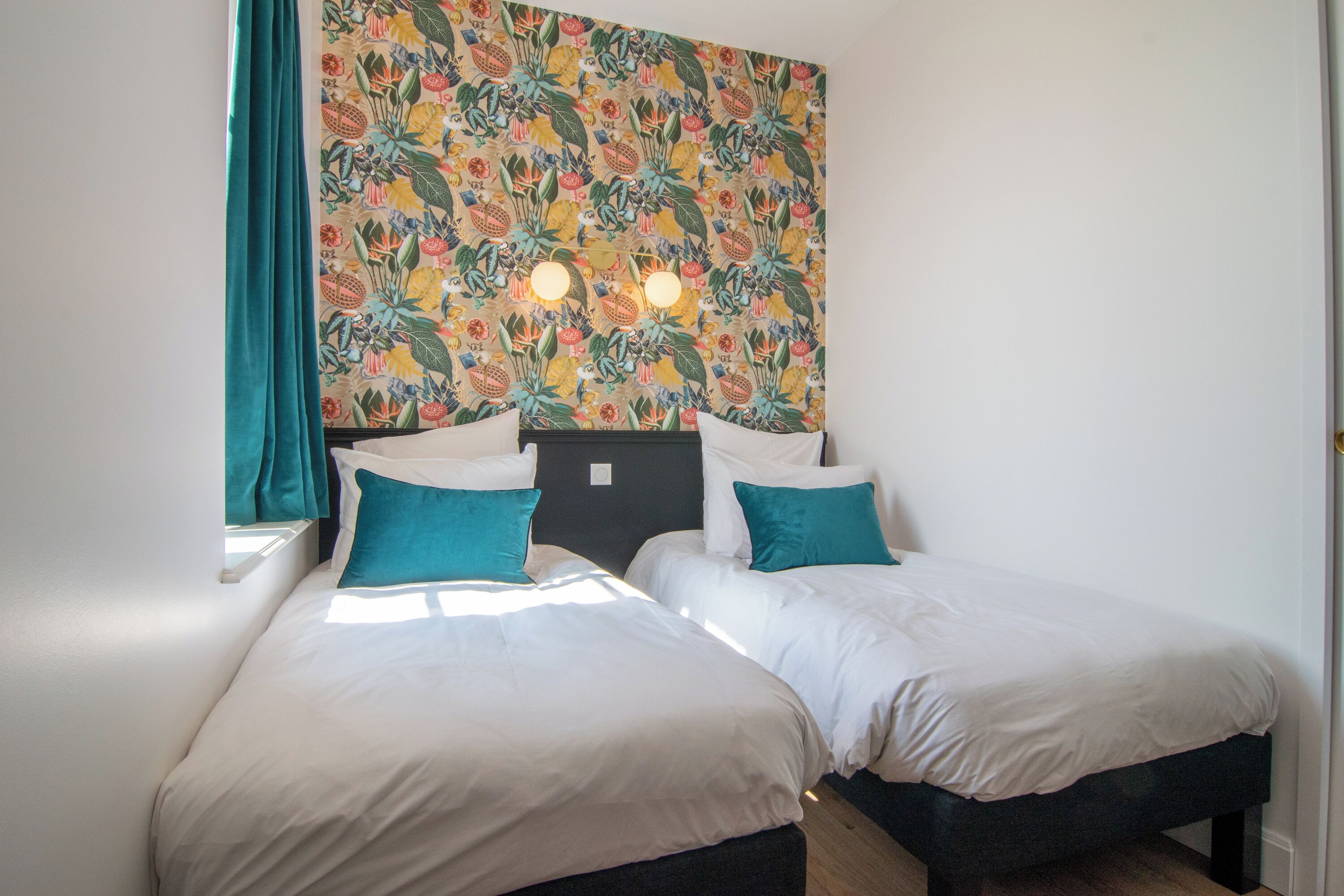 Ein Zimmer mit zwei Betten, türkisblauen Vorhängen und einer blumigen Wand.