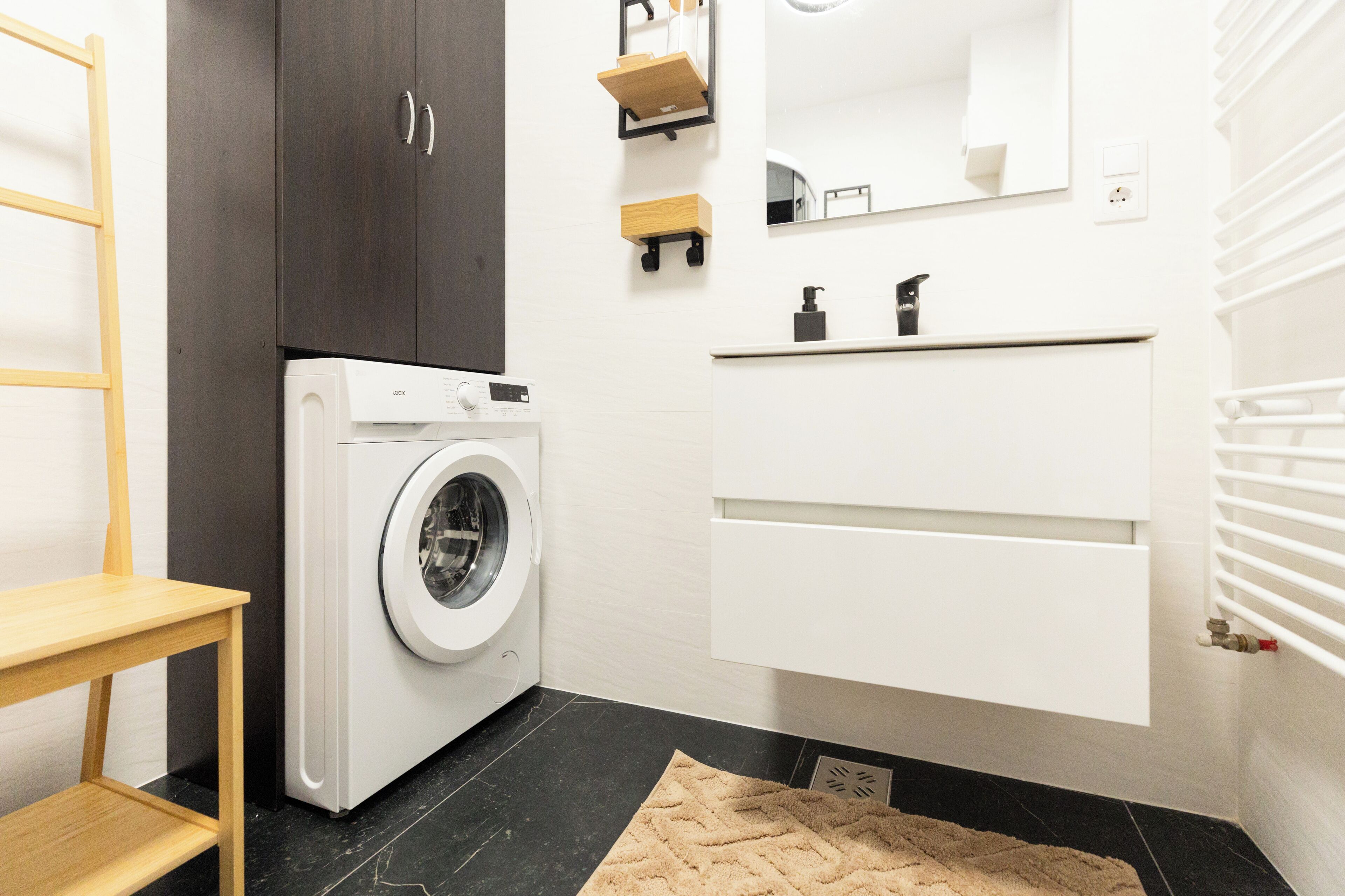 Een moderne wasruimte met een wasmachine, een houten plank, een spiegel en een wit kastje.