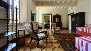 Ein geräumiges Zimmer mit schachigem Holzboden, einem großen Ohrensessel, einem gestreiften Sofa und einem Holzschrank.