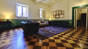 Ein geräumiges Wohnzimmer mit kariertem Holzboden, einer dunklen Couch und einem gemusterten Teppich. Zwei Fenster mit weißen Rahmen, ein an der Wand befestigter Bilderrahmen und eine grüne Tür sind zu sehen.