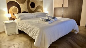 Una cama bien hecha con sábanas blancas y un cabecero de diseño circular. Sobre la cama hay toallas dobladas, y a la izquierda, una mesita de noche con una lámpara. El cuarto tiene suelo de madera y un armario de madera al fondo.