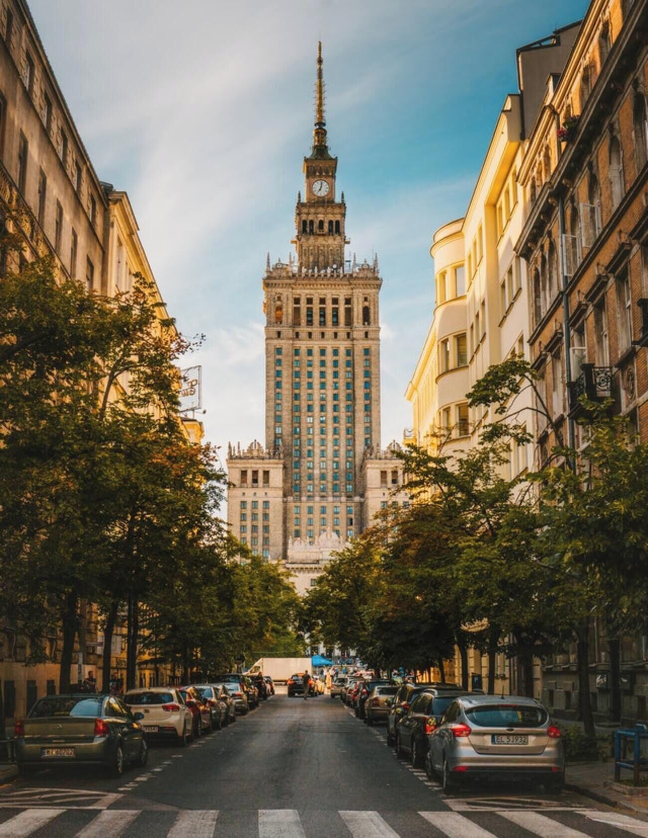 Une rue menant à un grand immeuble surmonté d’un clocher, bordée de voitures garées et d’arbres.