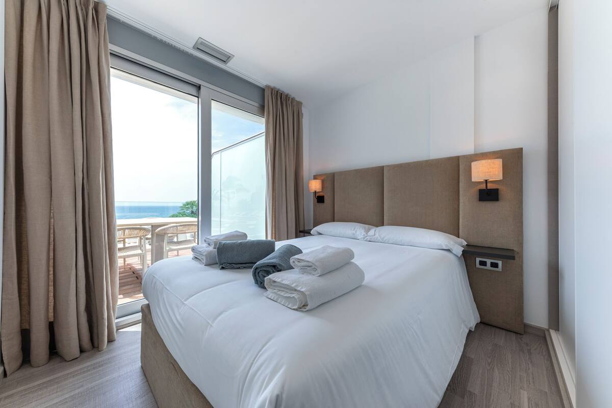 Une chambre d’hôtel avec un grand lit, deux lampes de chevet, une vue sur la mer et un balcon équipé d’une table et de chaises.