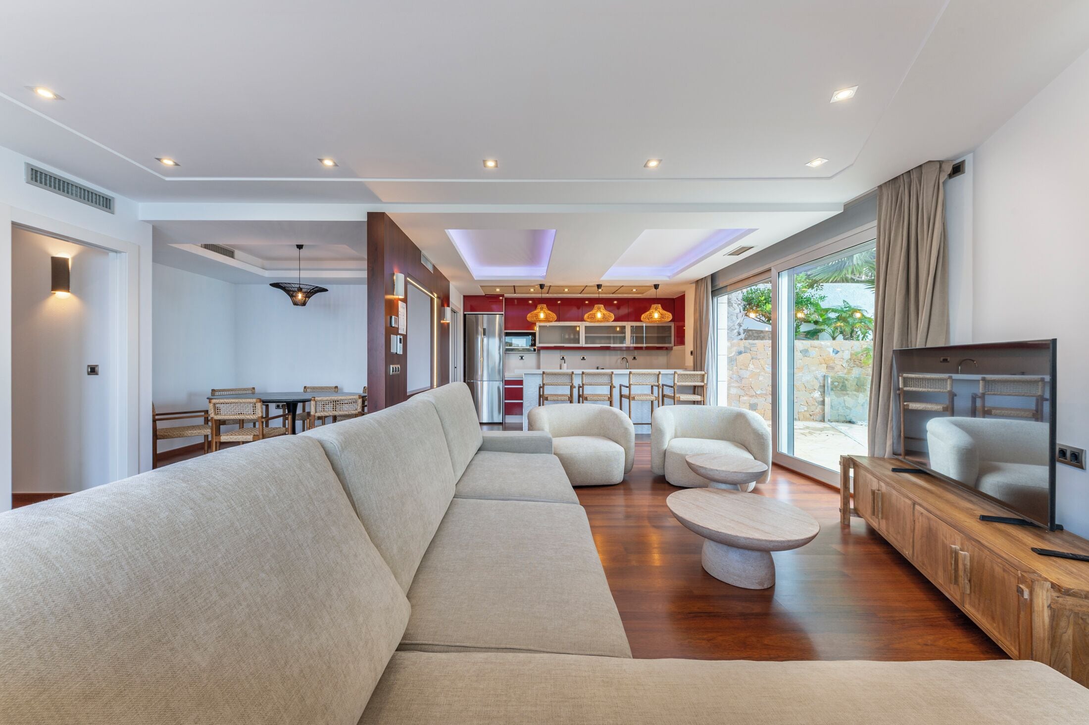Ein modernes Wohnzimmer mit einer großen beigen Couch, einem Holztisch und einem Fernseher auf einer Holzkommode. Im Hintergrund ist eine Küche mit roten Schränken und ein Essbereich mit Korbstühlen zu sehen.