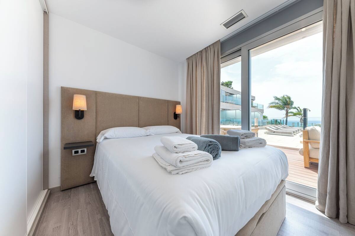 Une chambre d’hôtel avec un grand lit, deux lampes de chevet, une vue sur l’océan et un balcon équipé de chaises longues.