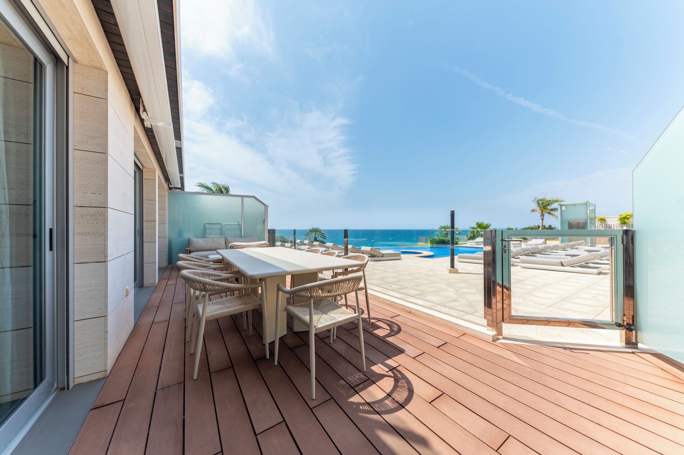 A wooden deck with a table and chairs, overlooking a pool and beach.