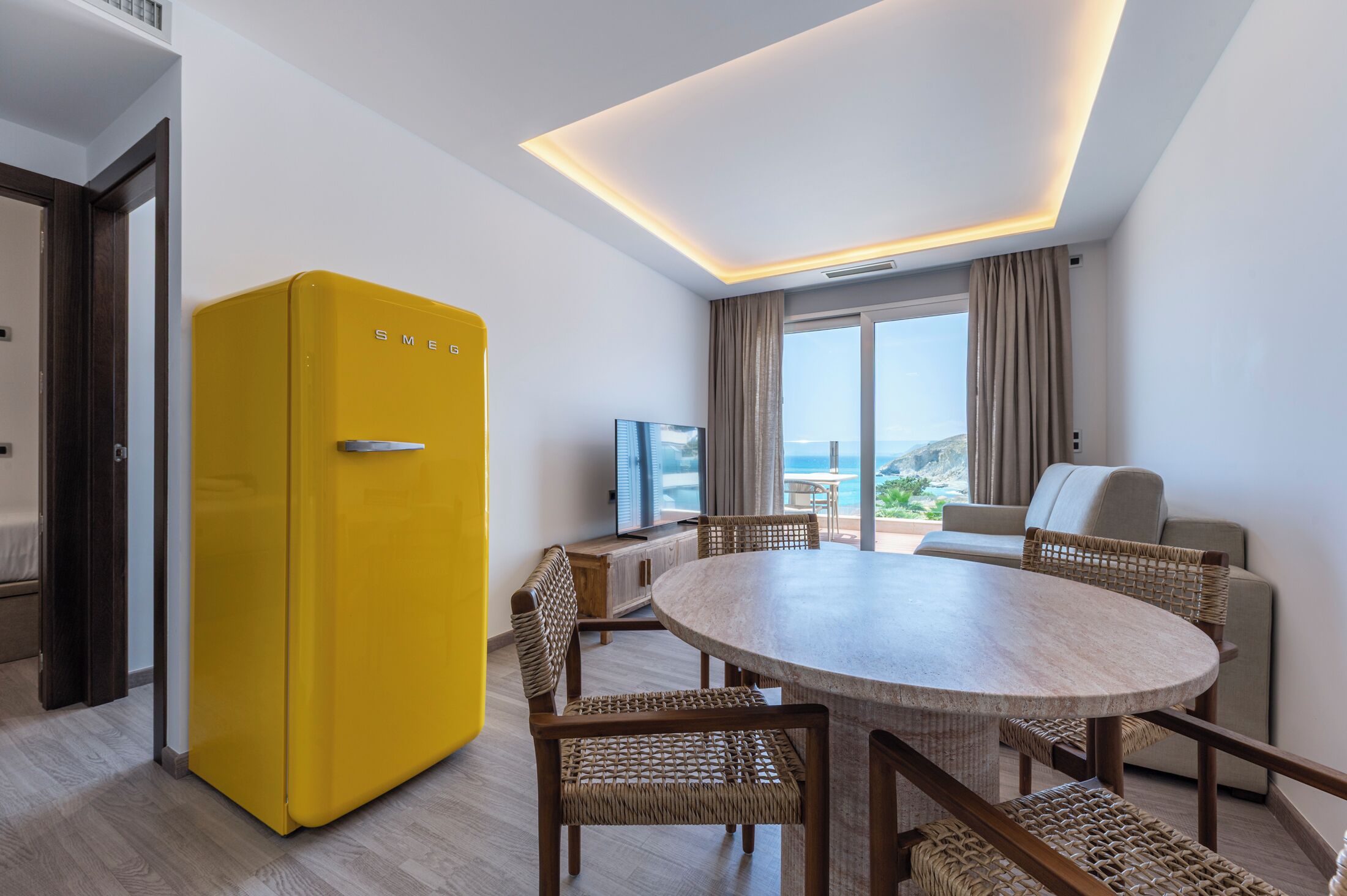 Habitación de hotel moderna con un refrigerador amarillo, una mesa de comedor redonda con sillas de mimbre, un sofá y un amplio ventanal con vistas al mar.