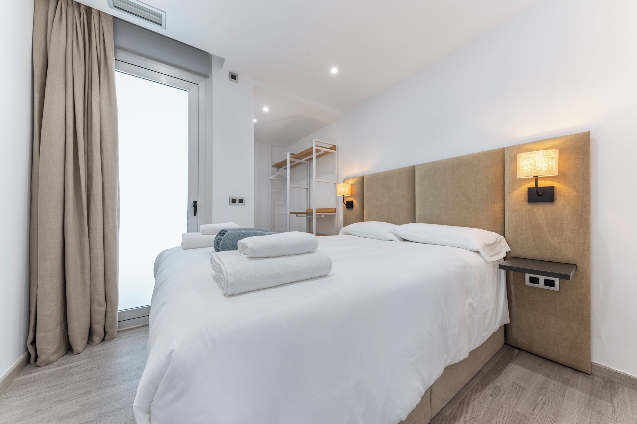 A modern hotel room with a large bed, a bedside table with a lamp, a wardrobe, and a view of the outside.