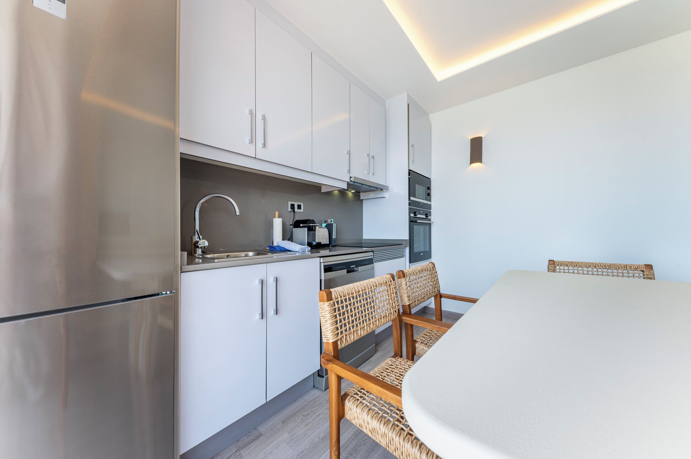 Una cocina moderna con alacenas blancas, electrodomésticos de acero inoxidable y un comedor con sillas de mimbre.