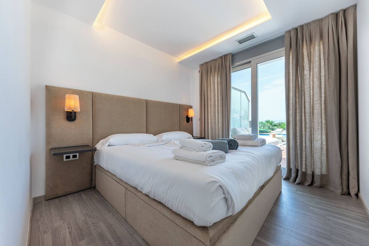 Une chambre d’hôtel moderne avec un grand lit, deux lampes de chevet, une vue sur la piscine et une fenêtre avec des rideaux.