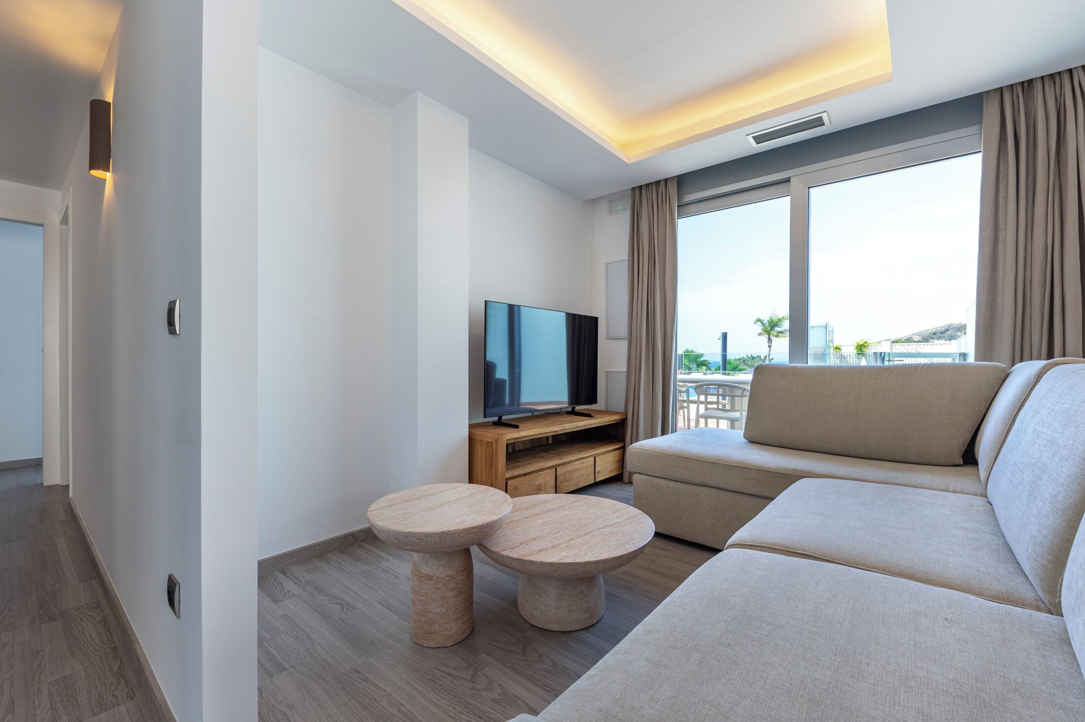 Una sala de estar moderna con un sofá, dos mesas redondas, un televisor sobre un mueble de madera y ventanas grandes con cortinas.