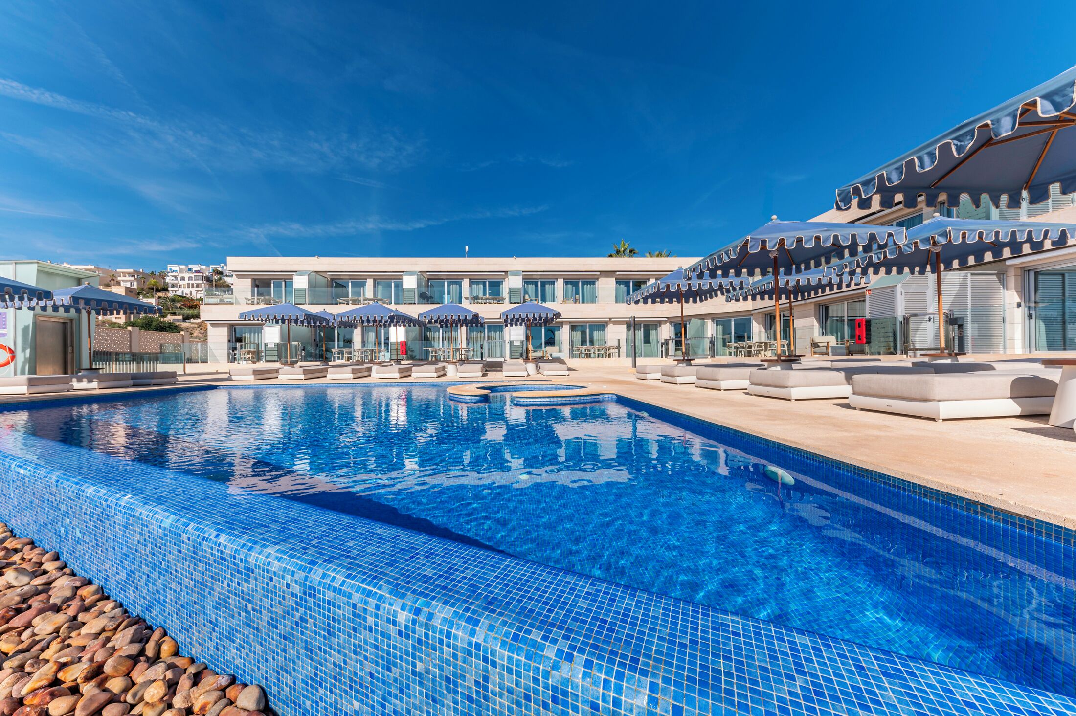 Una piscina con sillones y sombrillas, un edificio de diseño moderno y un cielo azul despejado.