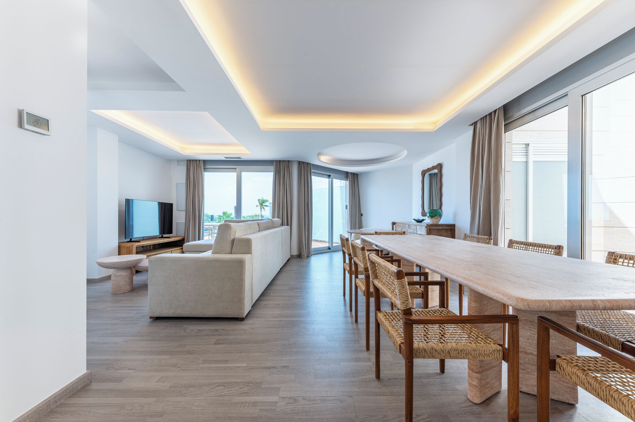 Una habitación de hotel moderna con una larga mesa de comedor, asientos cómodos, un televisor de pantalla plana y ventanas grandes que ofrecen vistas de la ciudad.