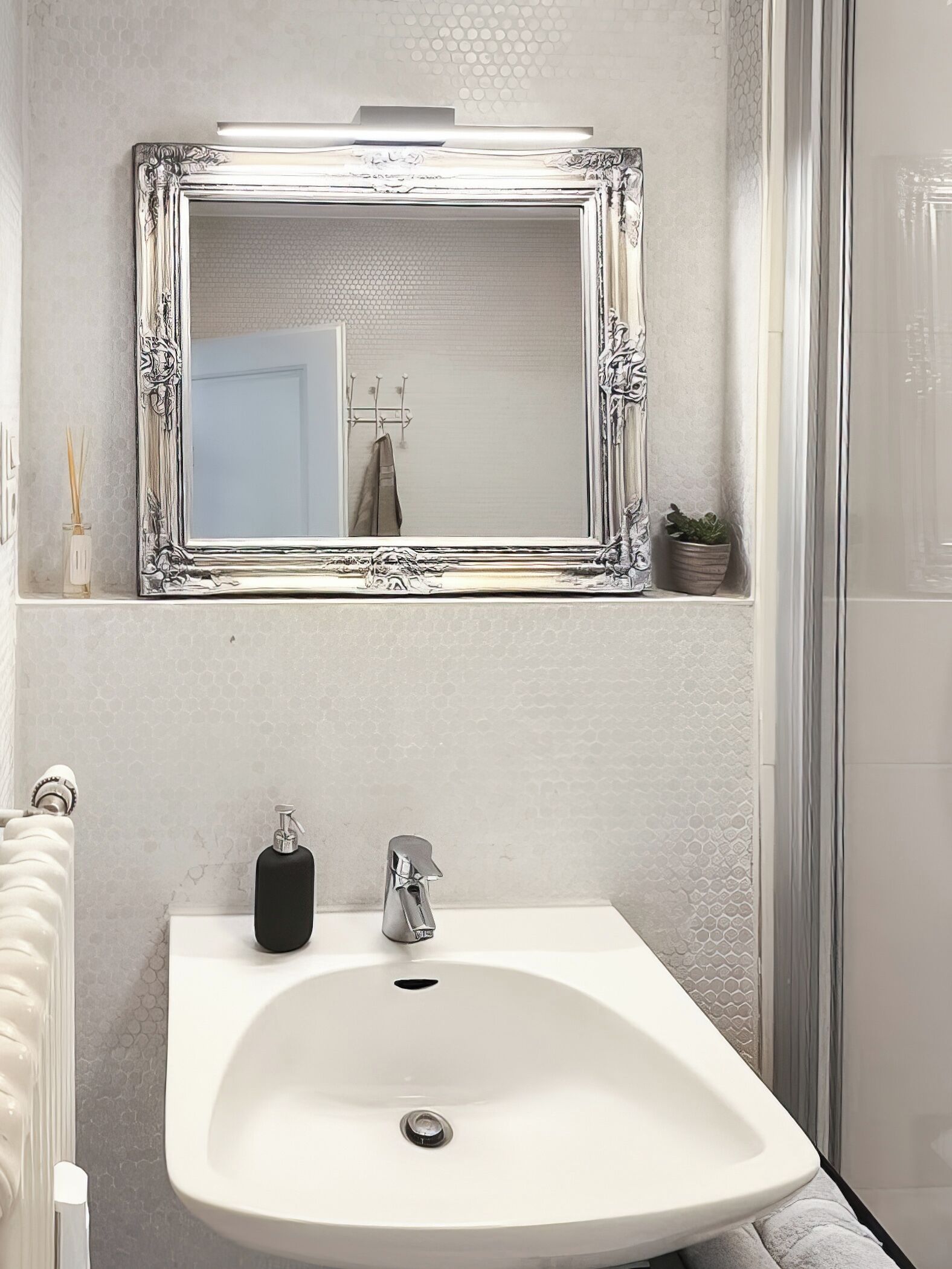 A bathroom with a white sink, a mirror with an ornate frame, a towel rack, a potted plant, and a shower area.