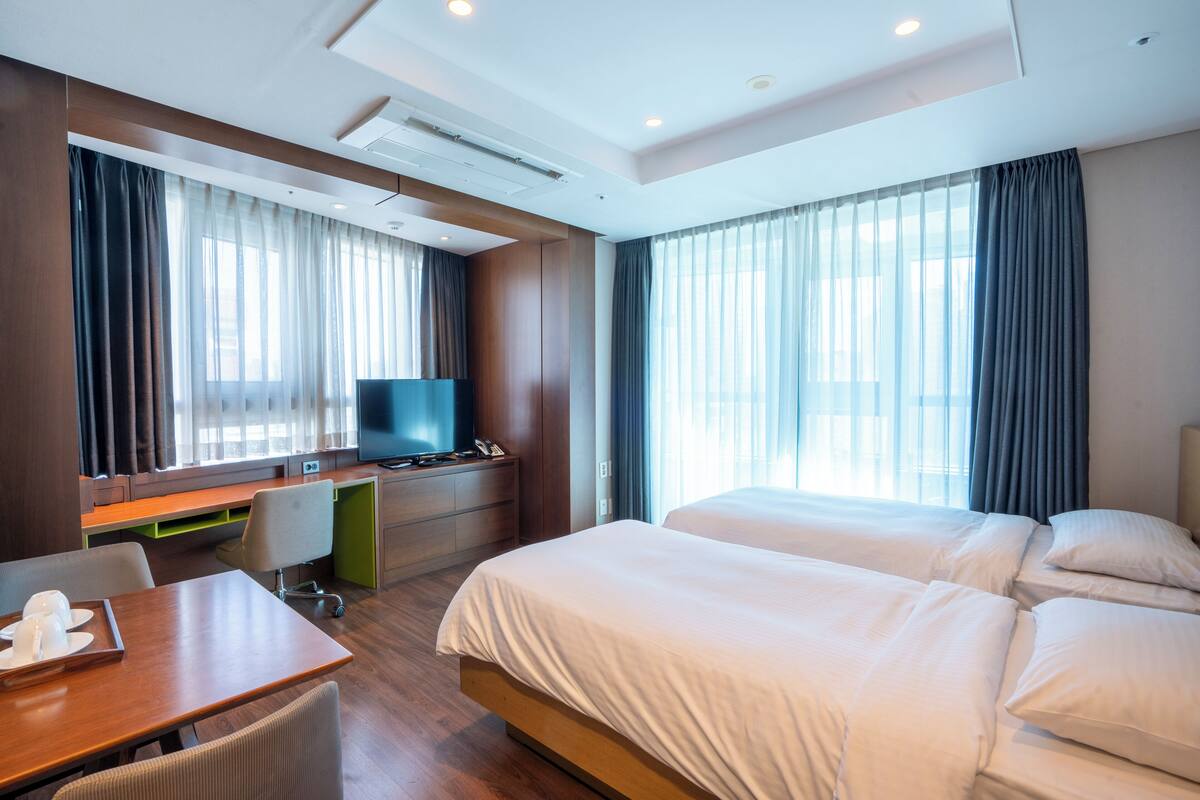 Habitación de hotel con cama, escritorio, silla, televisión y ventanas grandes con cortinas.