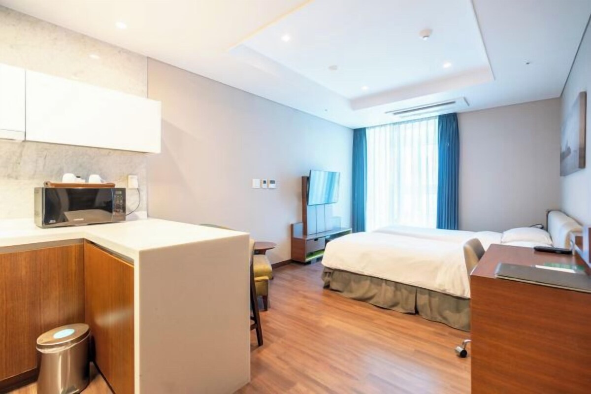 Une chambre d’hôtel équipée d’un lit, d’un bureau, d’une chaise, d’un four à micro-ondes, d’une poubelle et d’une fenêtre avec des rideaux.