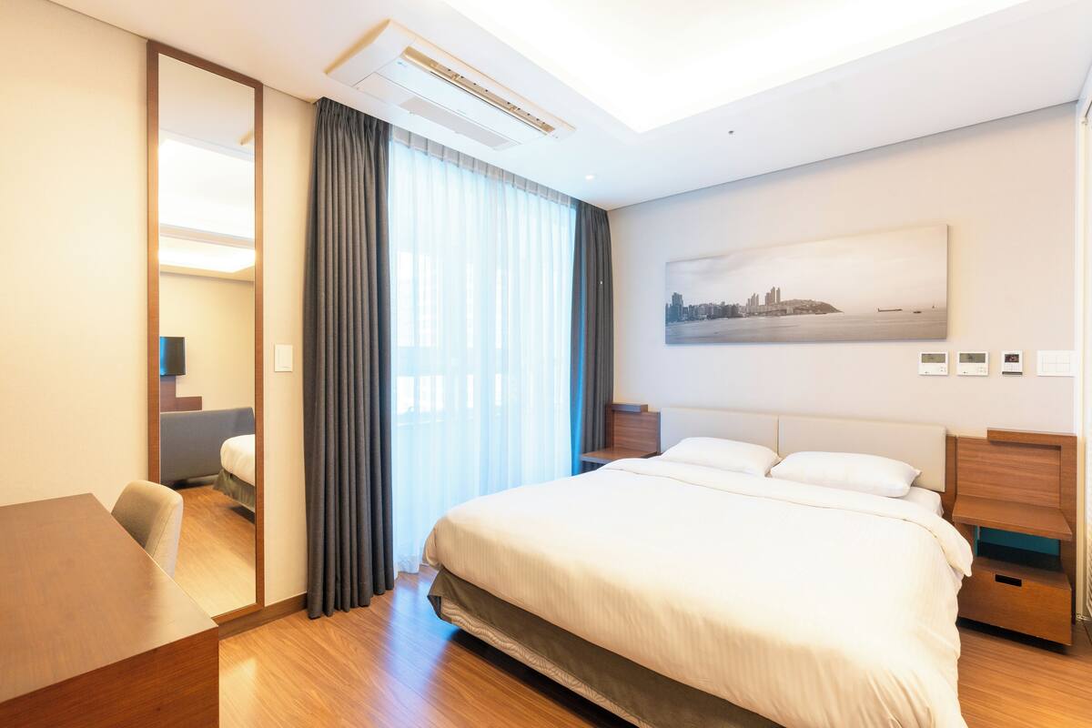 Un cuarto de hotel con una cama grande, un escritorio, una silla, un espejo, una mesita de noche y un cuadro de un paisaje urbano en la pared.