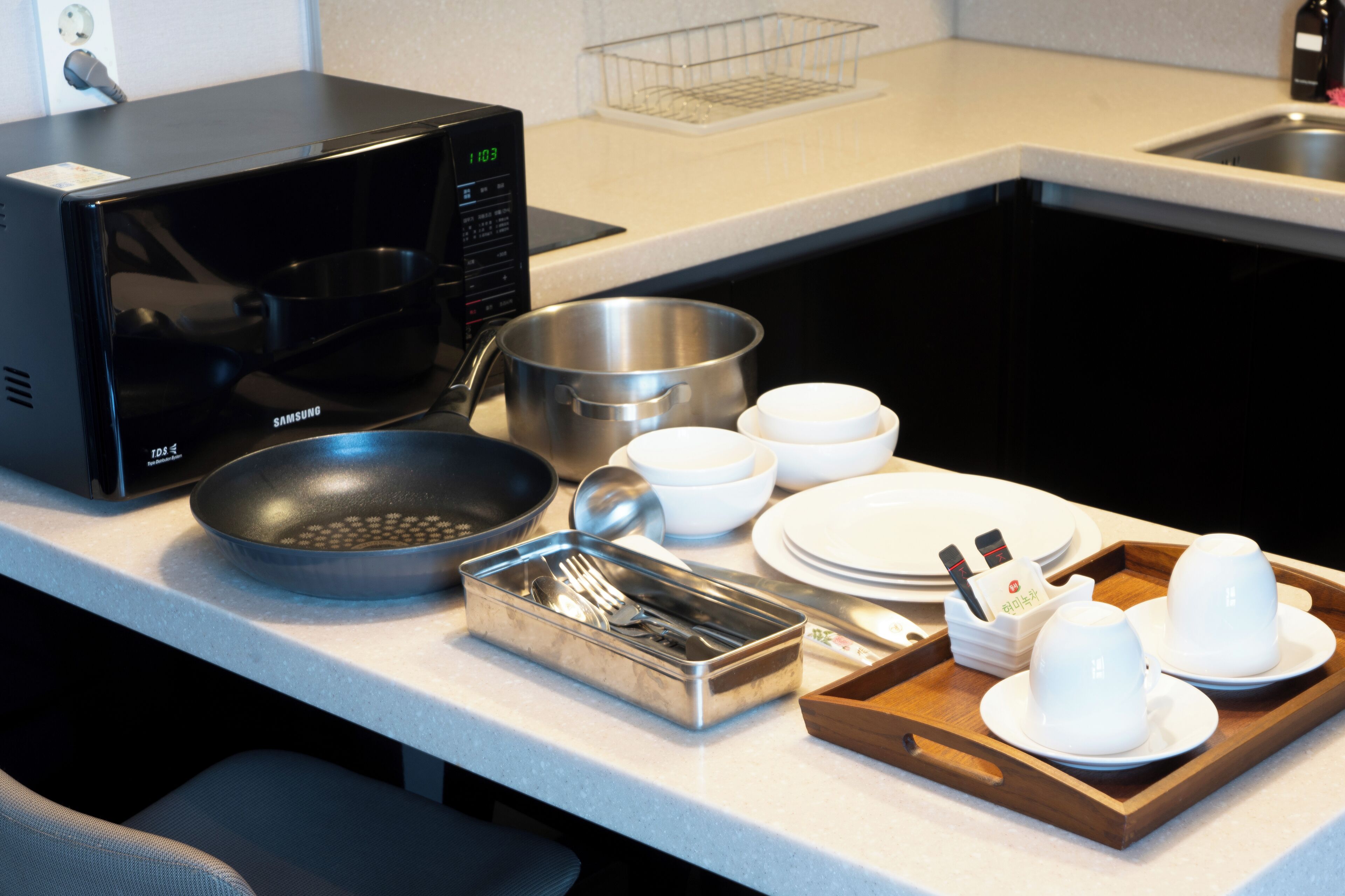 Una encimera de cocina con microondas, sartén, olla, cuencos, platos y una bandeja con tazas y utensilios.