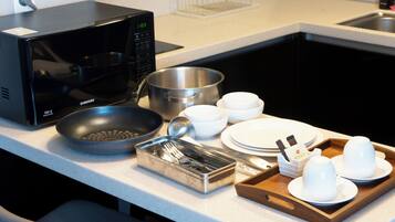 Una encimera de cocina con microondas, sartén, olla, cuencos, platos y una bandeja con tazas y utensilios.