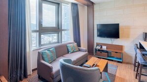 A modern living room with a sofa, armchair, coffee table, TV, and a city view through large windows. - hotellamouette (Busan)