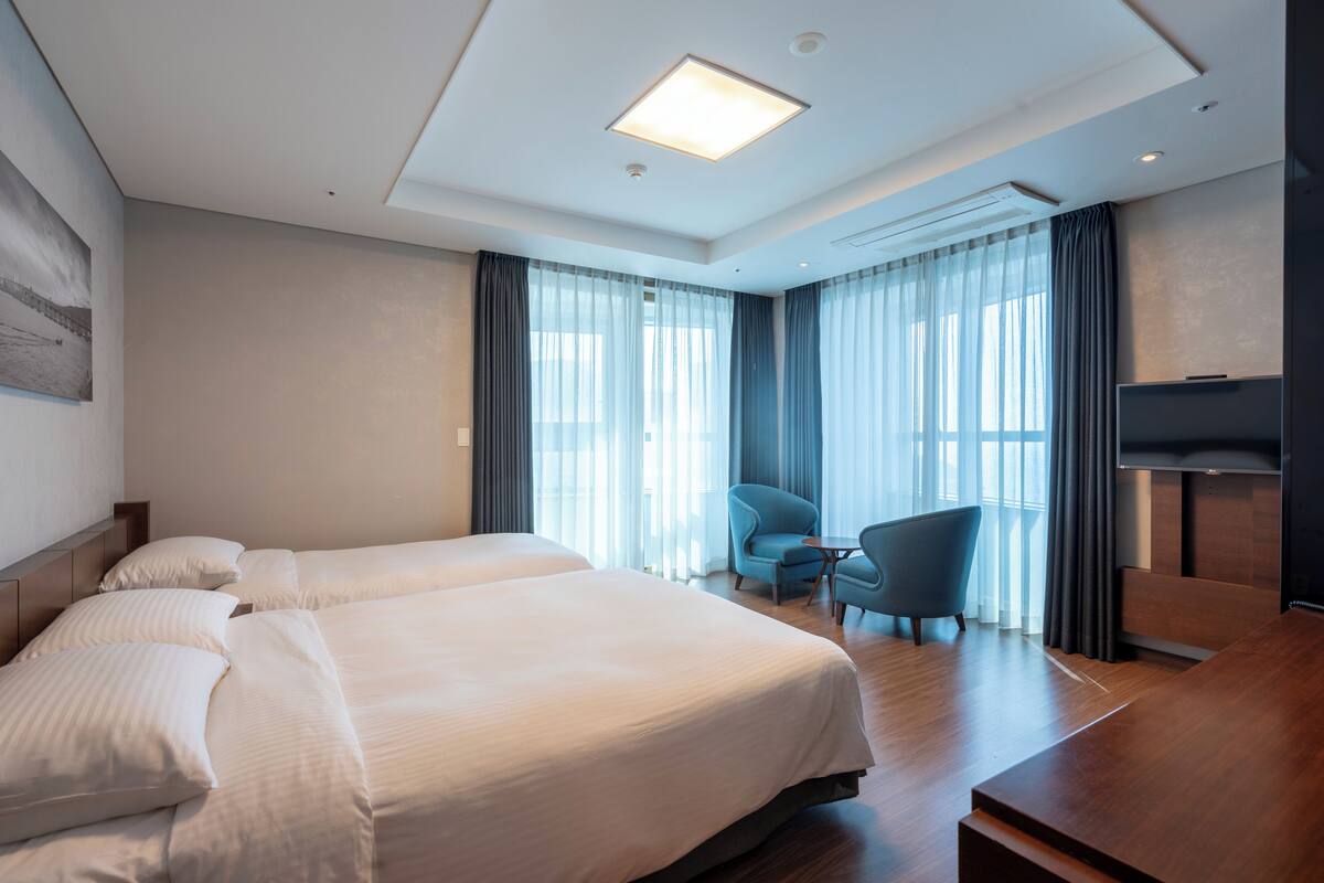 Habitación de hotel con dos camas, un escritorio, una silla, un televisor y ventanas grandes con cortinas.