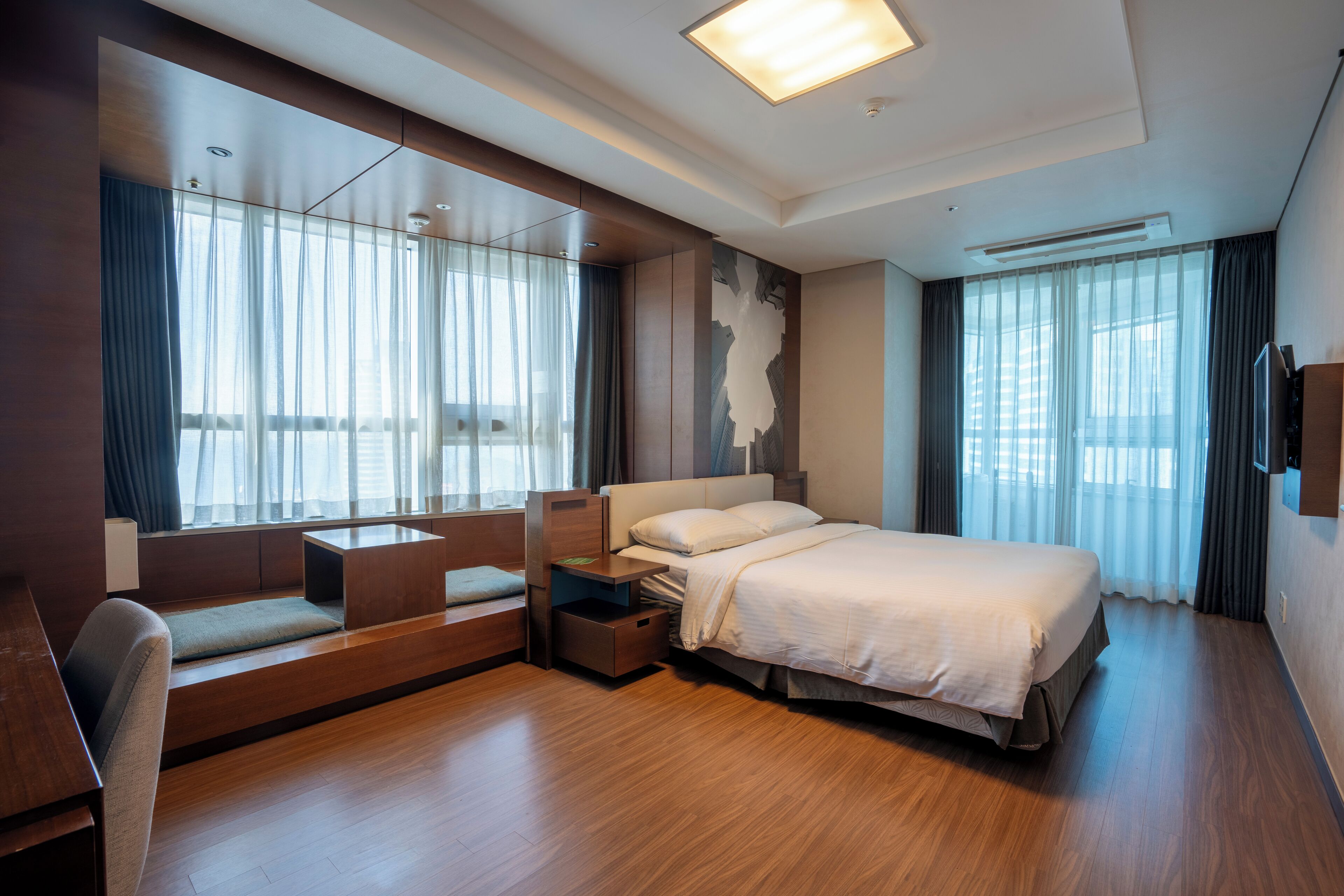 A modern hotel room with a large bed, a seating area with a bench and a small table, a flat-screen TV mounted on the wall, and large windows with curtains.