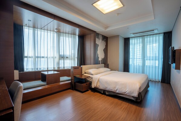 A modern hotel room with a large bed, a seating area with a bench and a small table, a flat-screen TV mounted on the wall, and large windows with curtains. - hotellamouette (Busan)