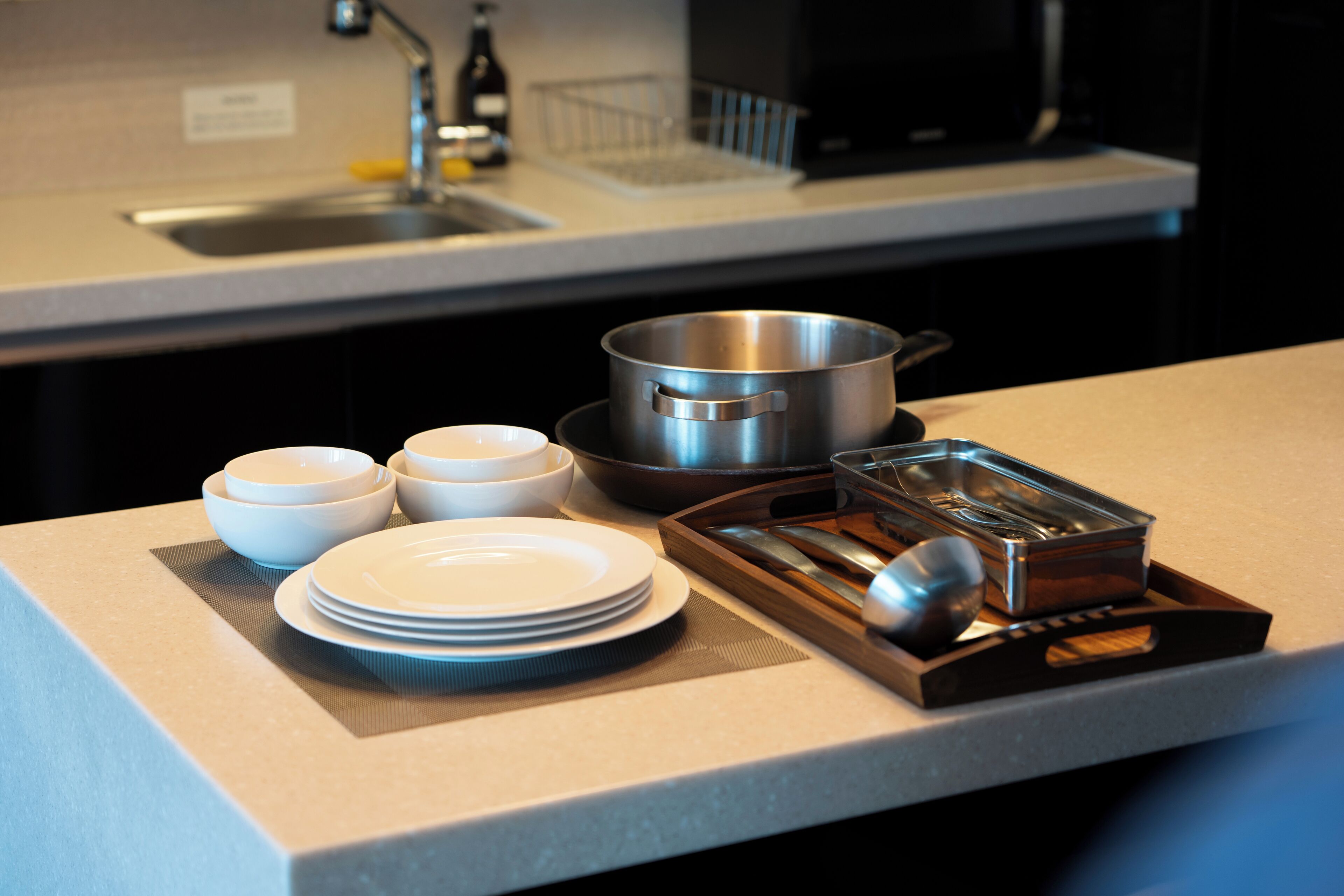 A kitchen counter with stacked white plates, a metal pot, a wooden tray with utensils, and a metal container.